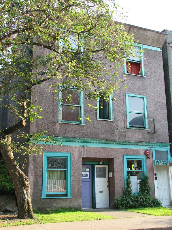 A photo of an old apartment building with dirty pink-beige exterior walls, window frames painted turquoise blue, and a budding tree out front